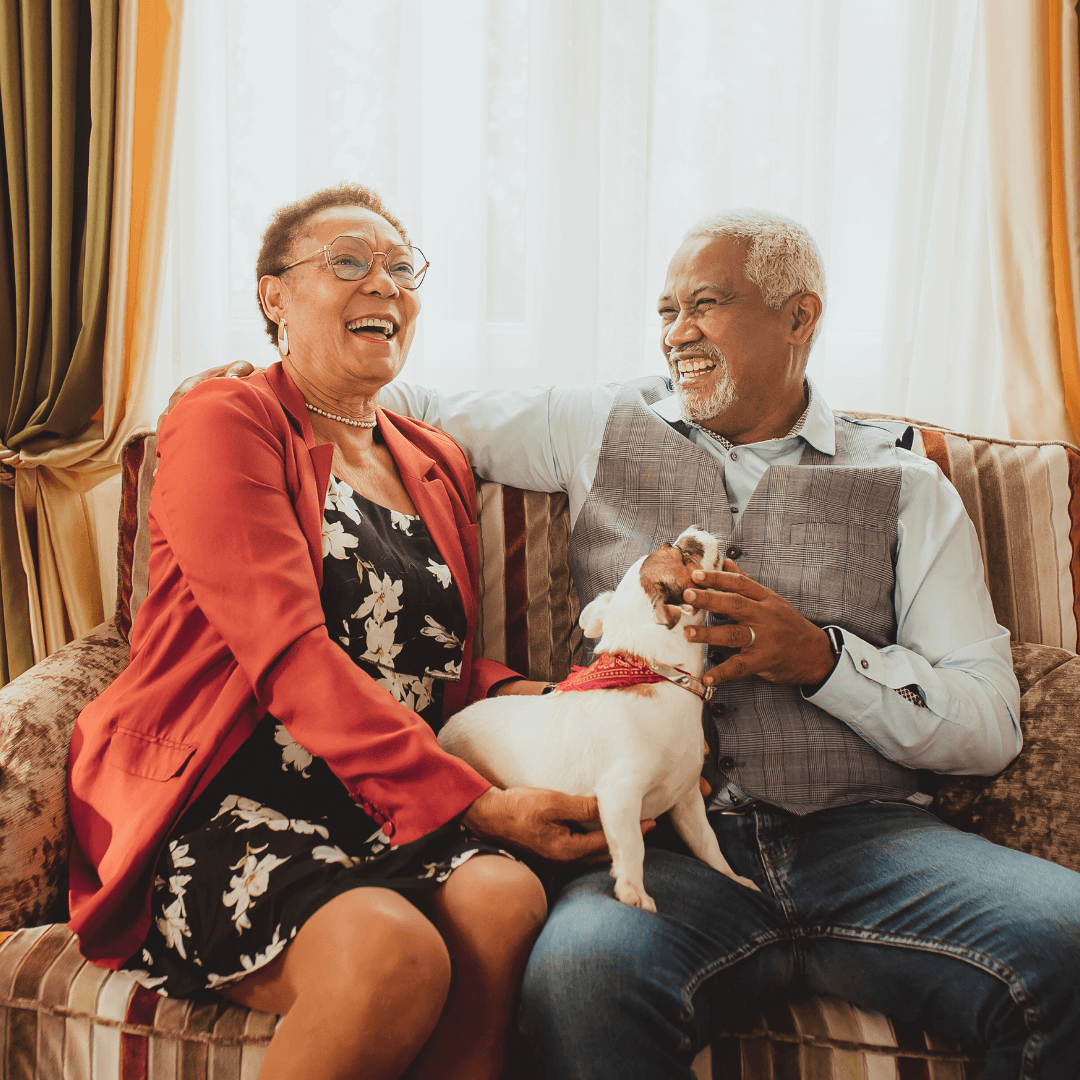 Senior couple sitting together on a couch, laughing and petting their dog.